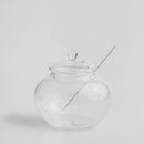 Transparentná sklenená nádoba na med s pokrývkou a lyžicou.